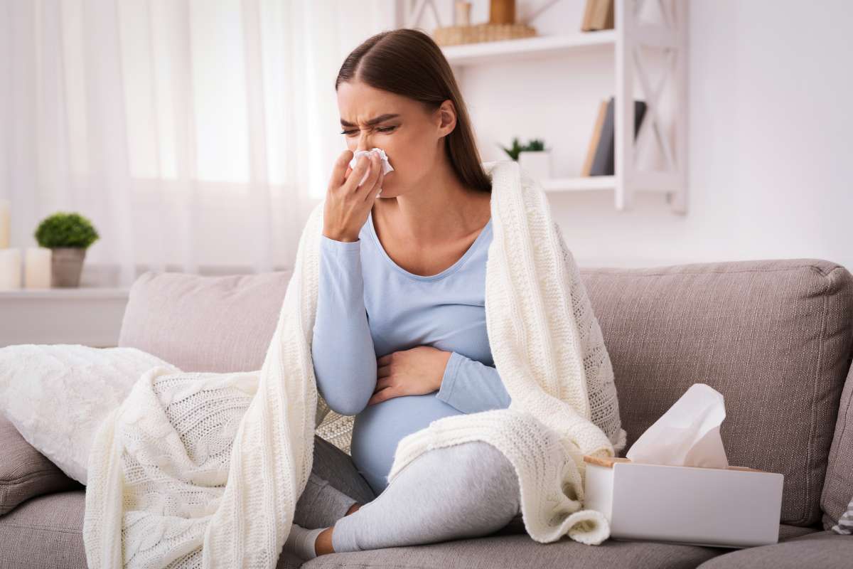 Pregnant woman blowing nose in tissue sitting on sofa indoor- Infections During Pregnancy