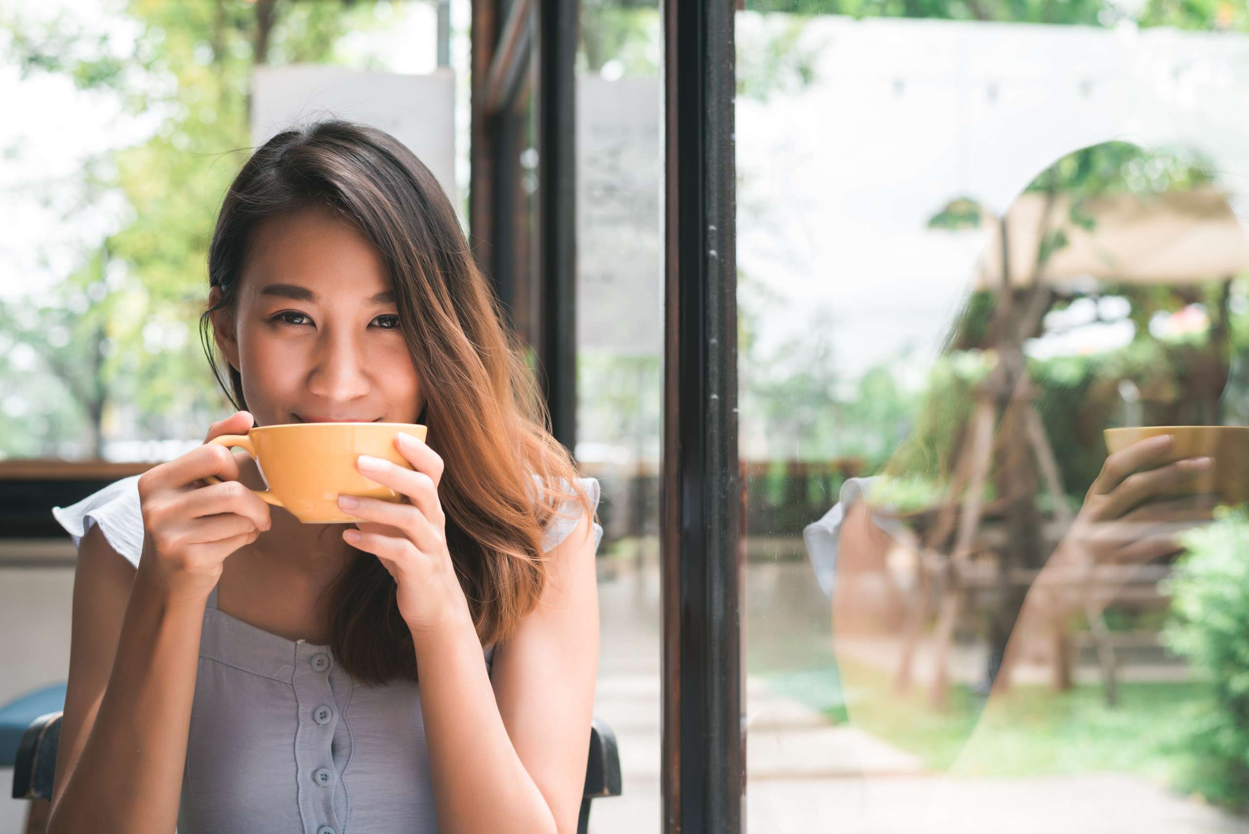 Cheerful asian young woman drinking warm coffee or tea enjoying it while sitting in cafe. Attractive happy asian woman holding a cup of coffee.