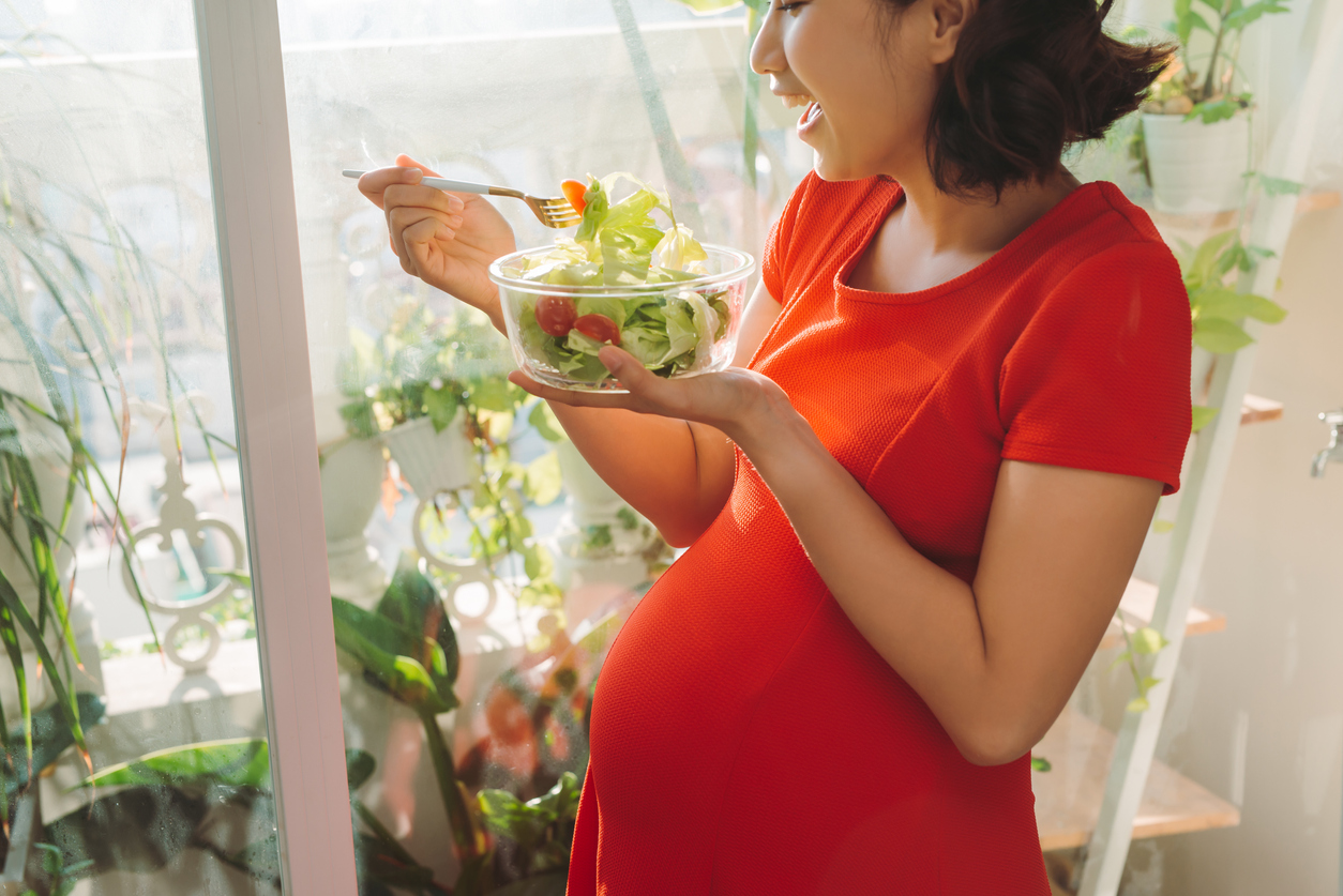 Full of vitamins. Energetic good looking pregnant woman eating her meat while carrying plate in a hand and relaxing against the window
