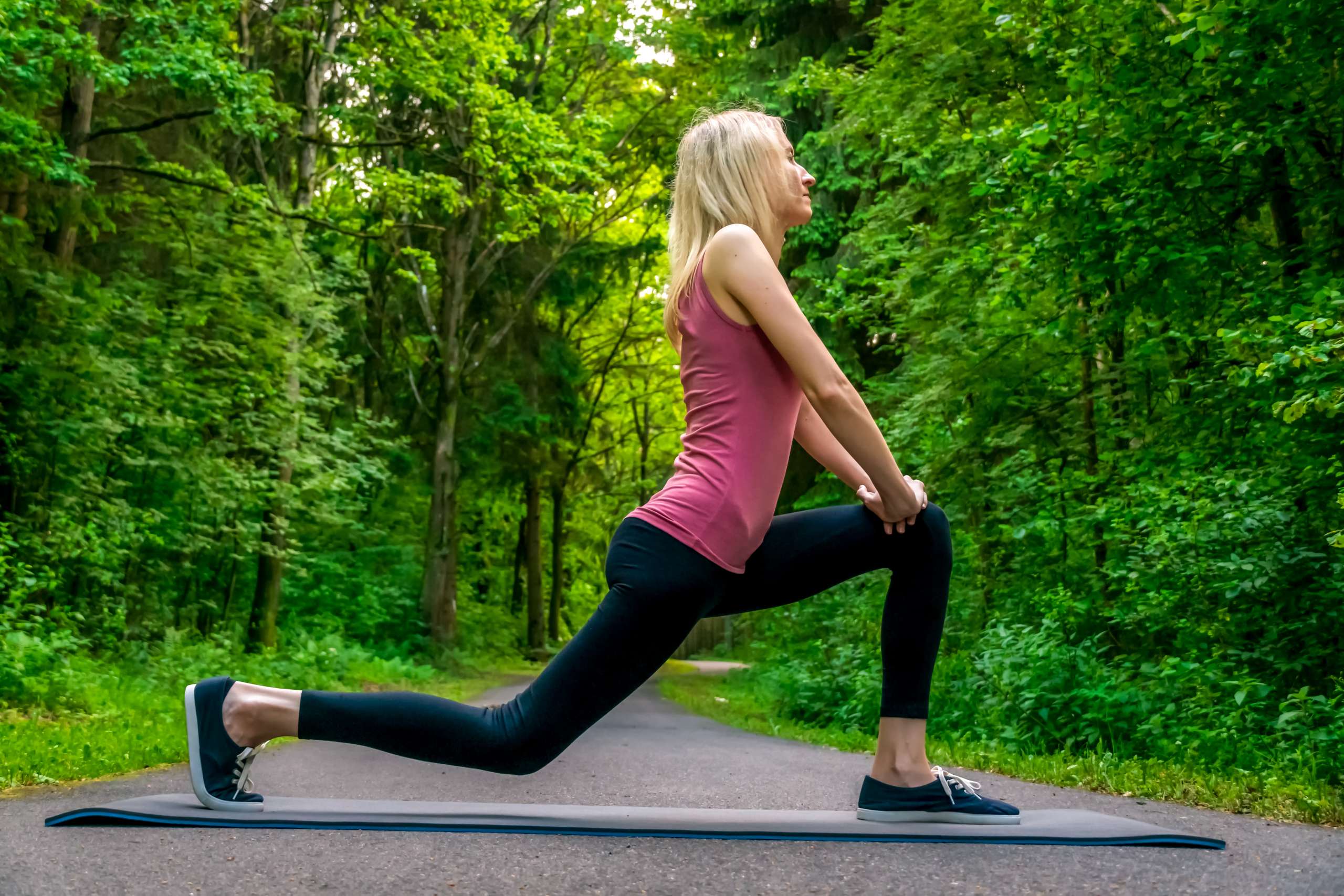 young blonde woman does a leg lunge exercise in the woods on a summer day