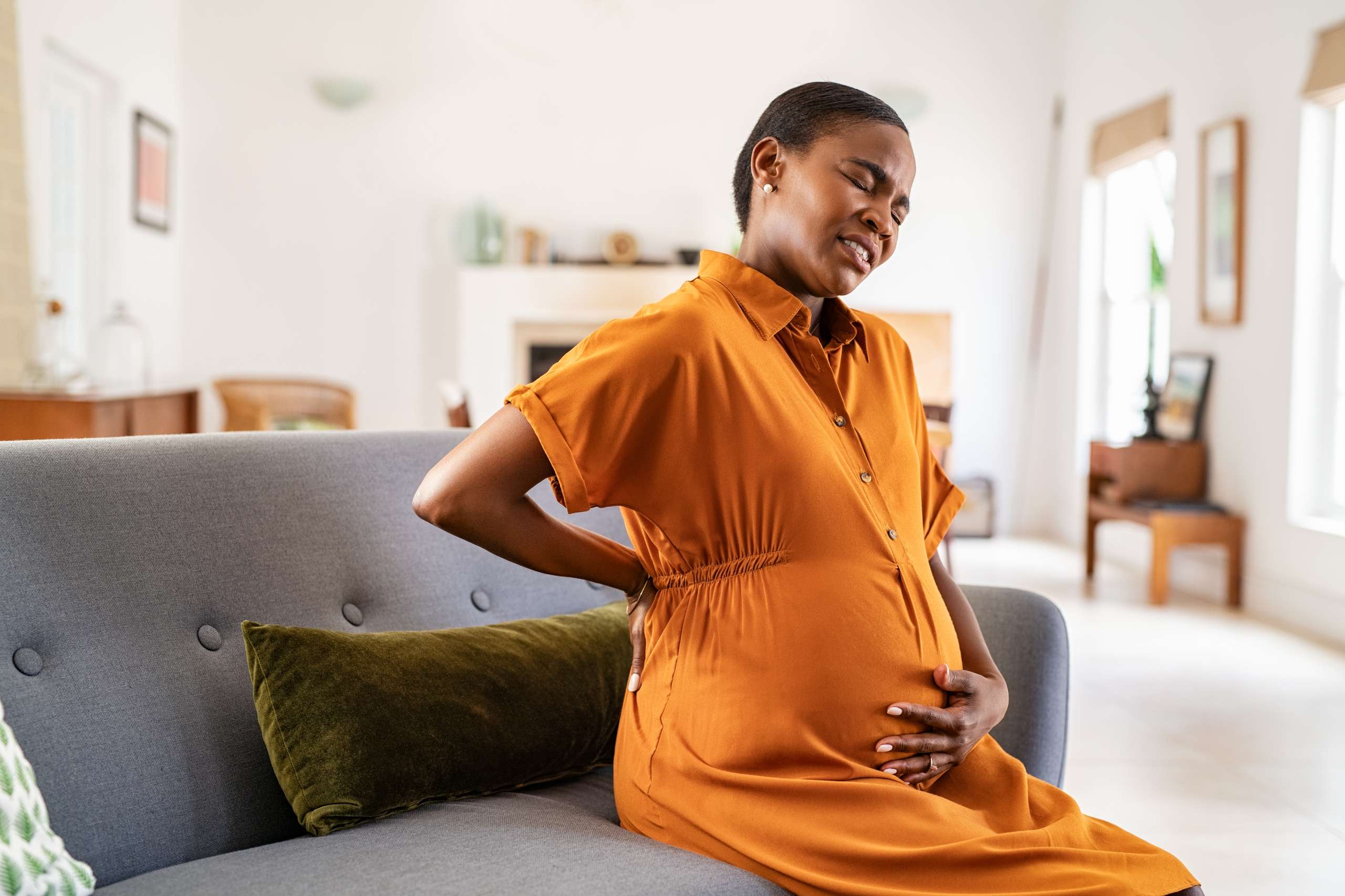 Mature pregnant woman having a backache while relaxing on couch at home. Unhappy black expecting woman suffering from lower back pain sitting on sofa with copy space. Tired middle aged african pregnant woman suffer from lower back pain on last month of pregnancy - Third Trimester Complications