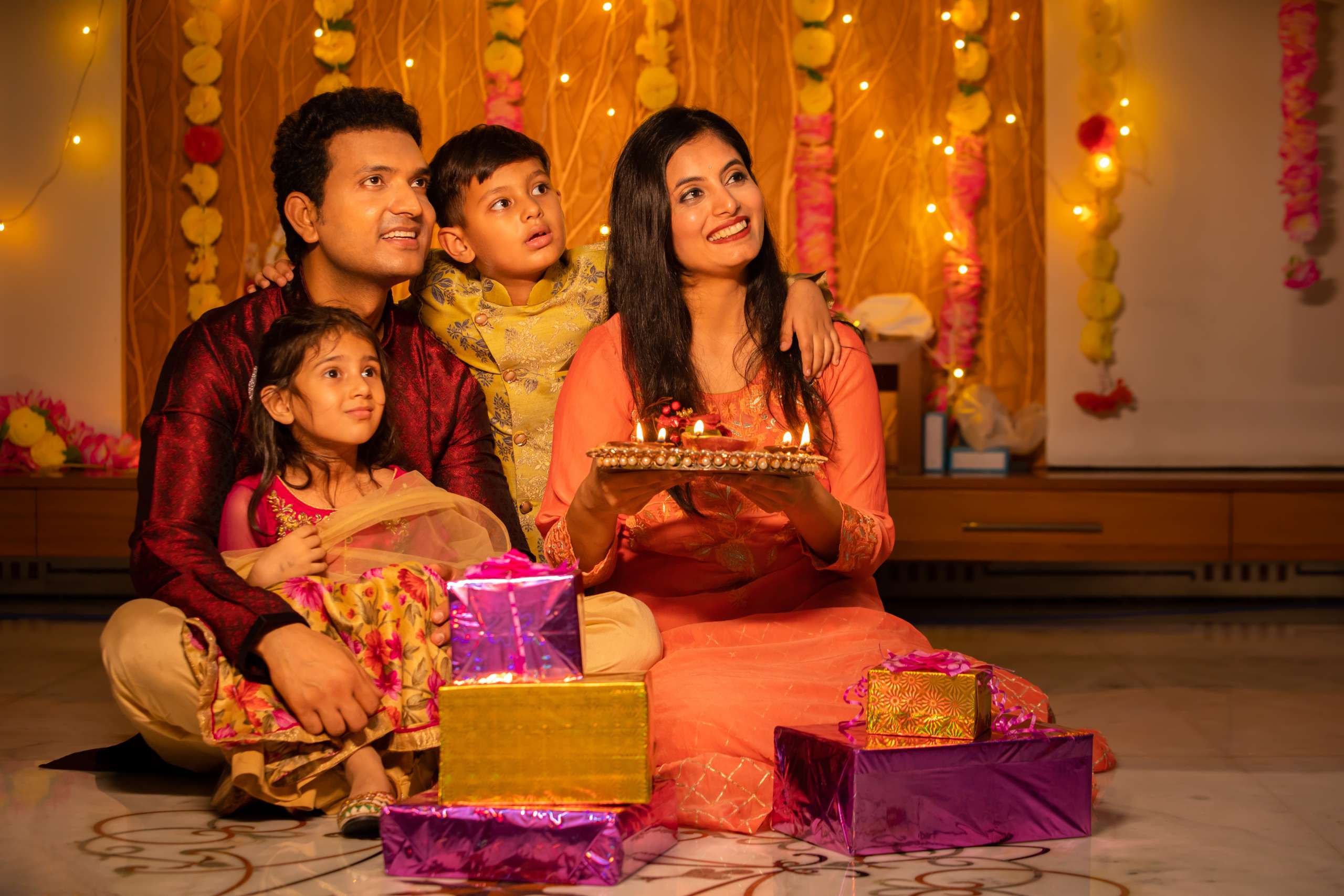 Happy young Indian family in traditional dress with lots of gifts around sitting on floor celebrating diwali festival at home.